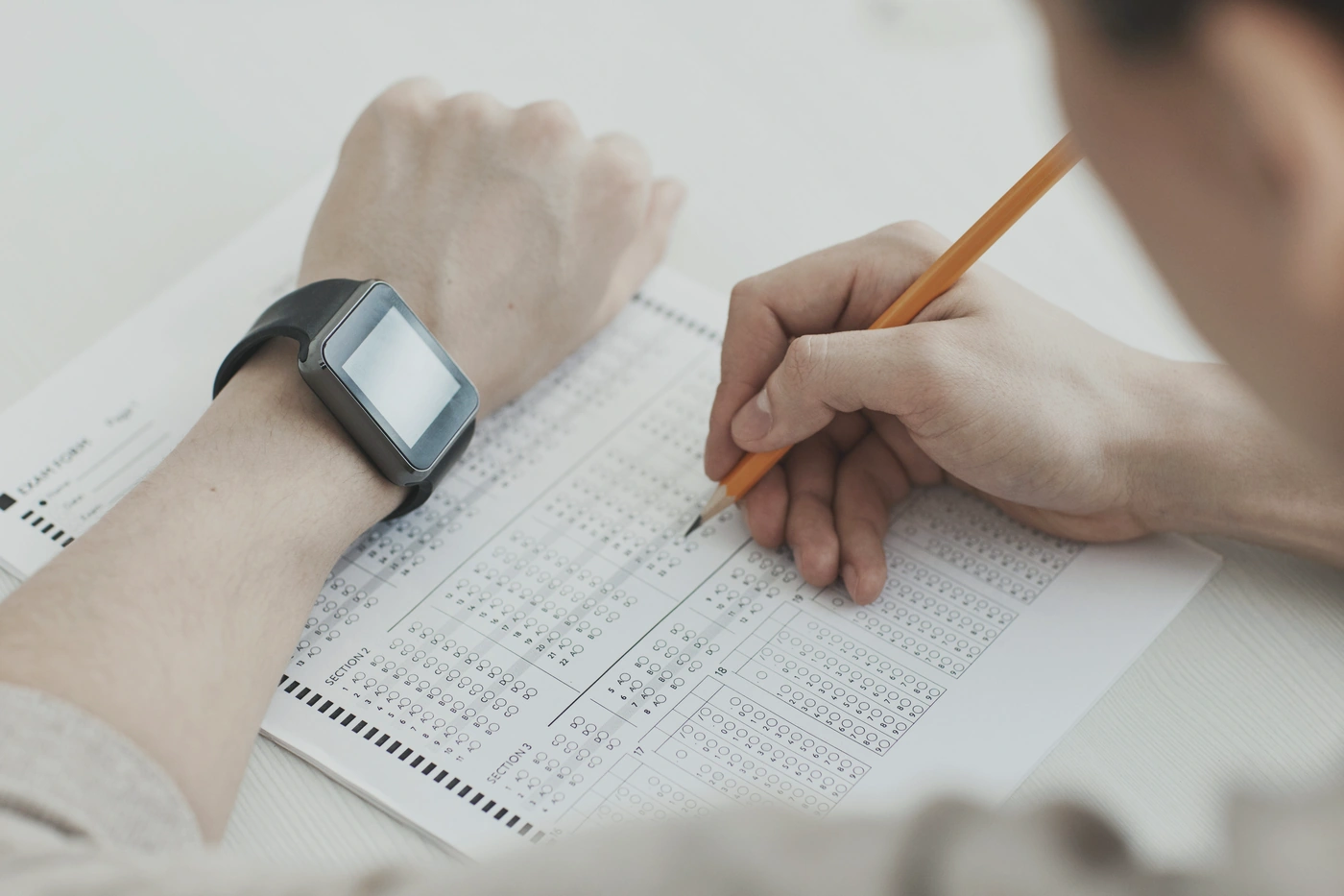 Close-up of two hands at a desk, one holding an orange pencil and filling in a multiple-choice bubble answer sheet.